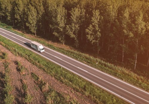 Aerial view of lorry truck on the road through green forest landscape in summer afternoon, drone pov high angle view photography