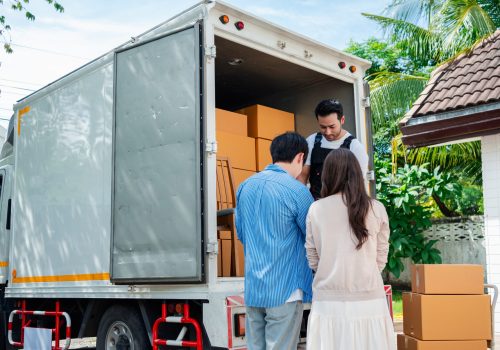 Asian Couple check while unloading boxes and furniture from a pickup truck to a new house with service cargo two men movers worker in uniform lifting boxes. concept of Home moving and delivery.