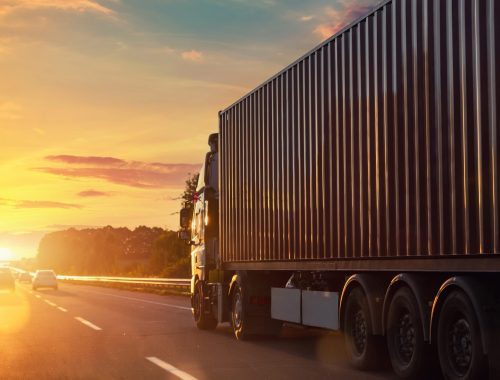 Scenic back view big long heavy semi-treailer truck with sea shipping container driving highway dramatic warm morning evening sunrise sun sky sunset. Cargo transport industry background concept.